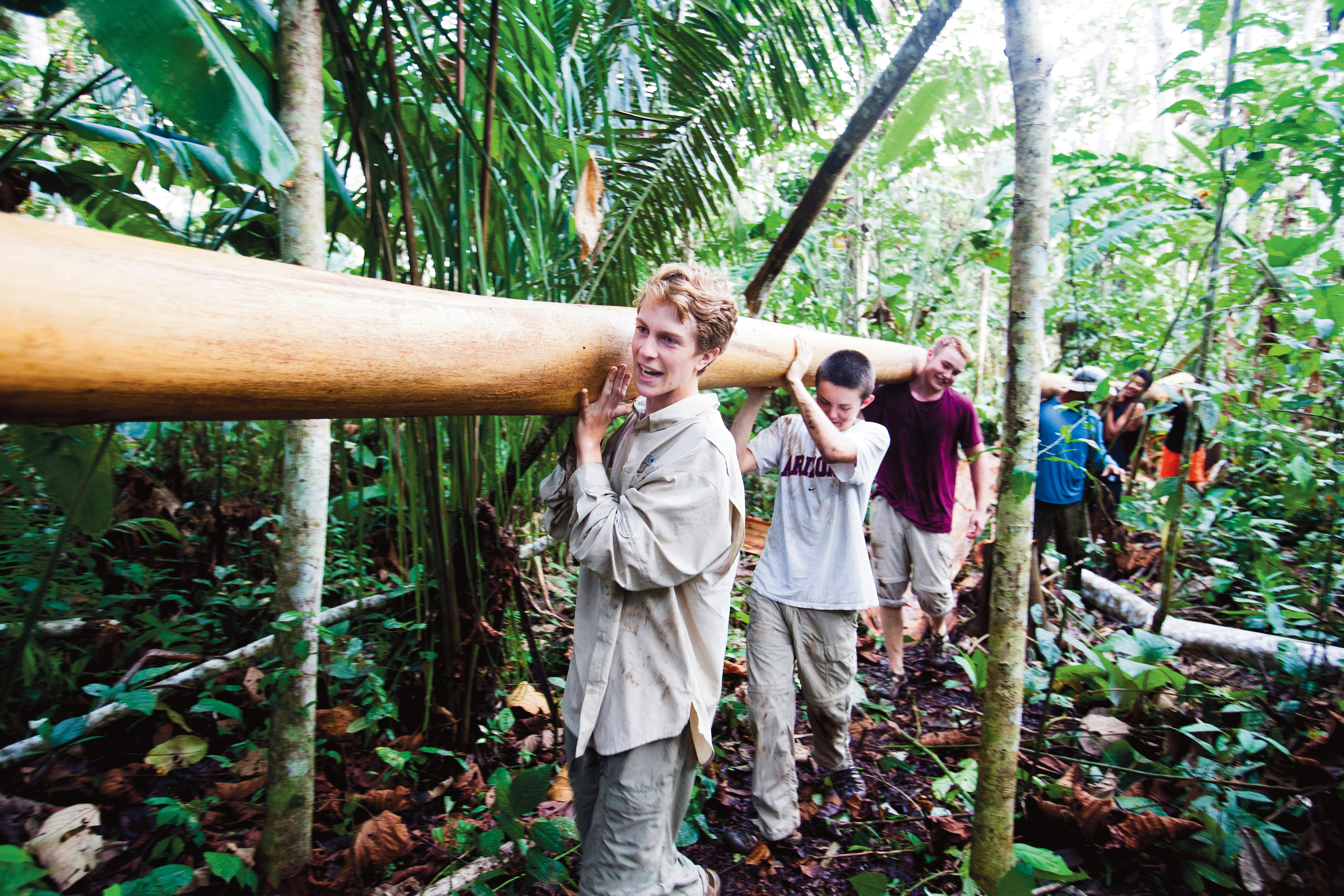 Group of young men carrying a large log through a dense forest.