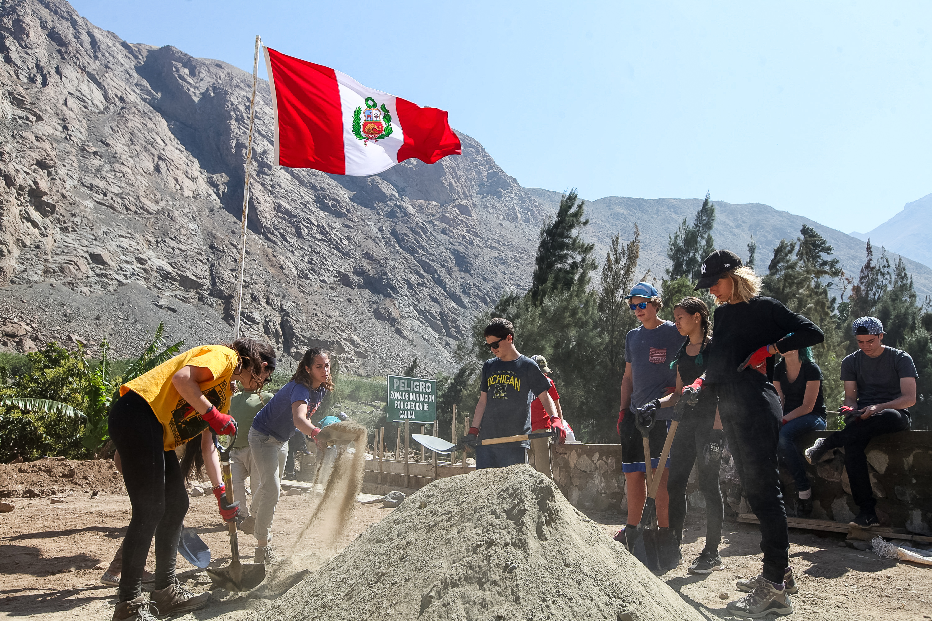Students shovel sand into a pile with the Peruvian flag and mountains behind them