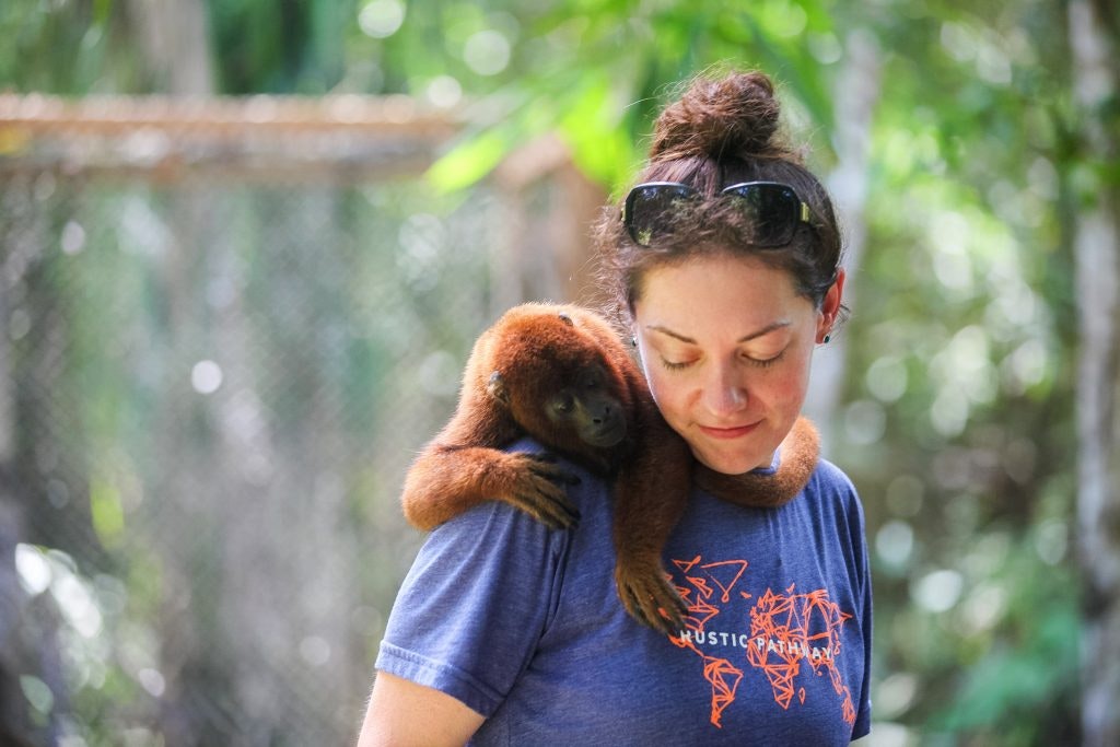 Howler monkey resting on the shoulder of a person wearing a Rustic Pathways shirt in a rainforest environment.