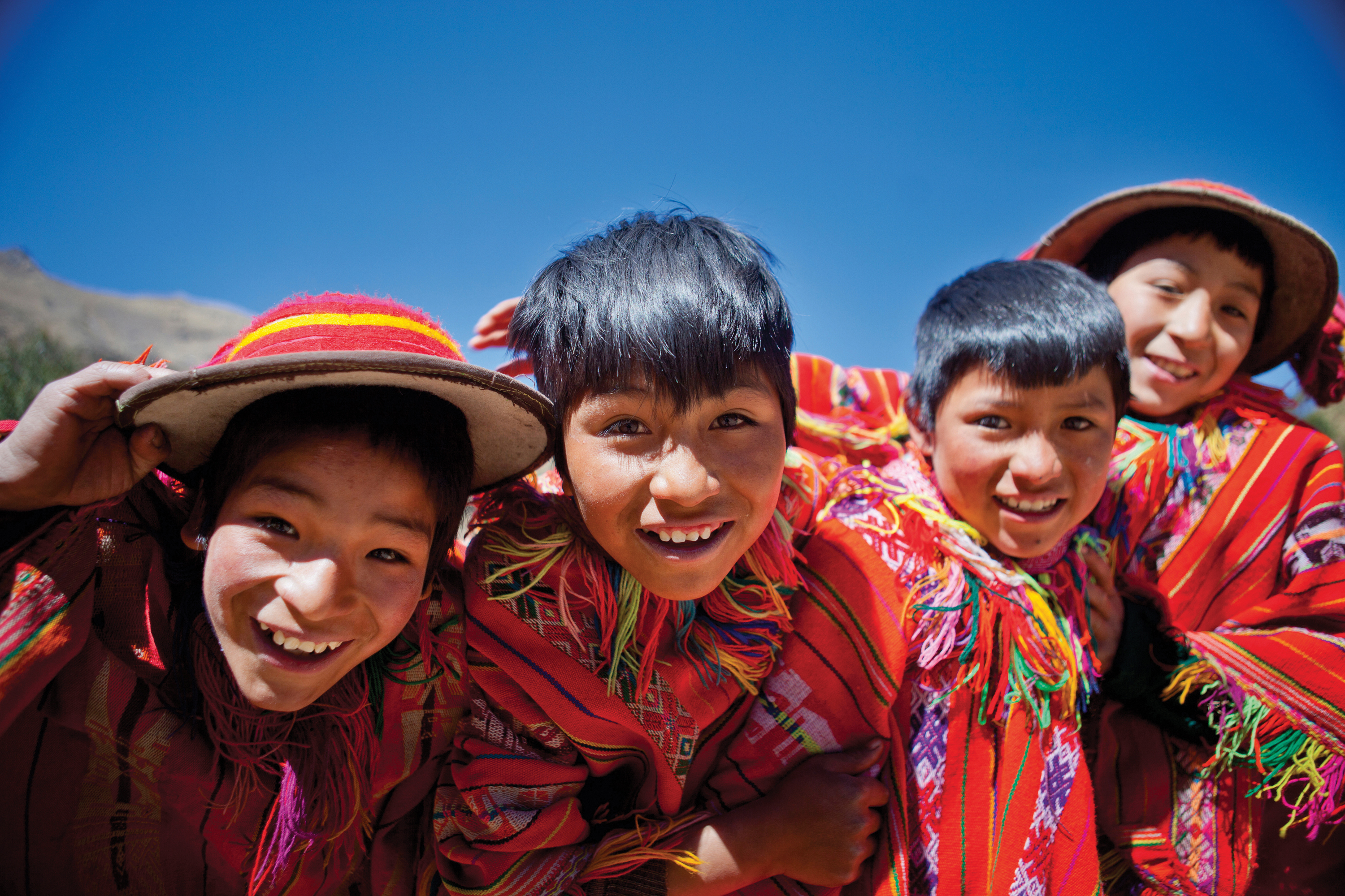 Local boys in traditional red Andean clothing and hats smile against a bright blue sky