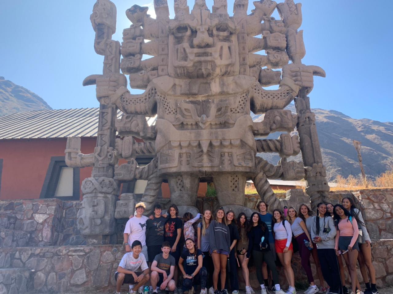 Students pose beneath a large carved stone monument with mountains in the background