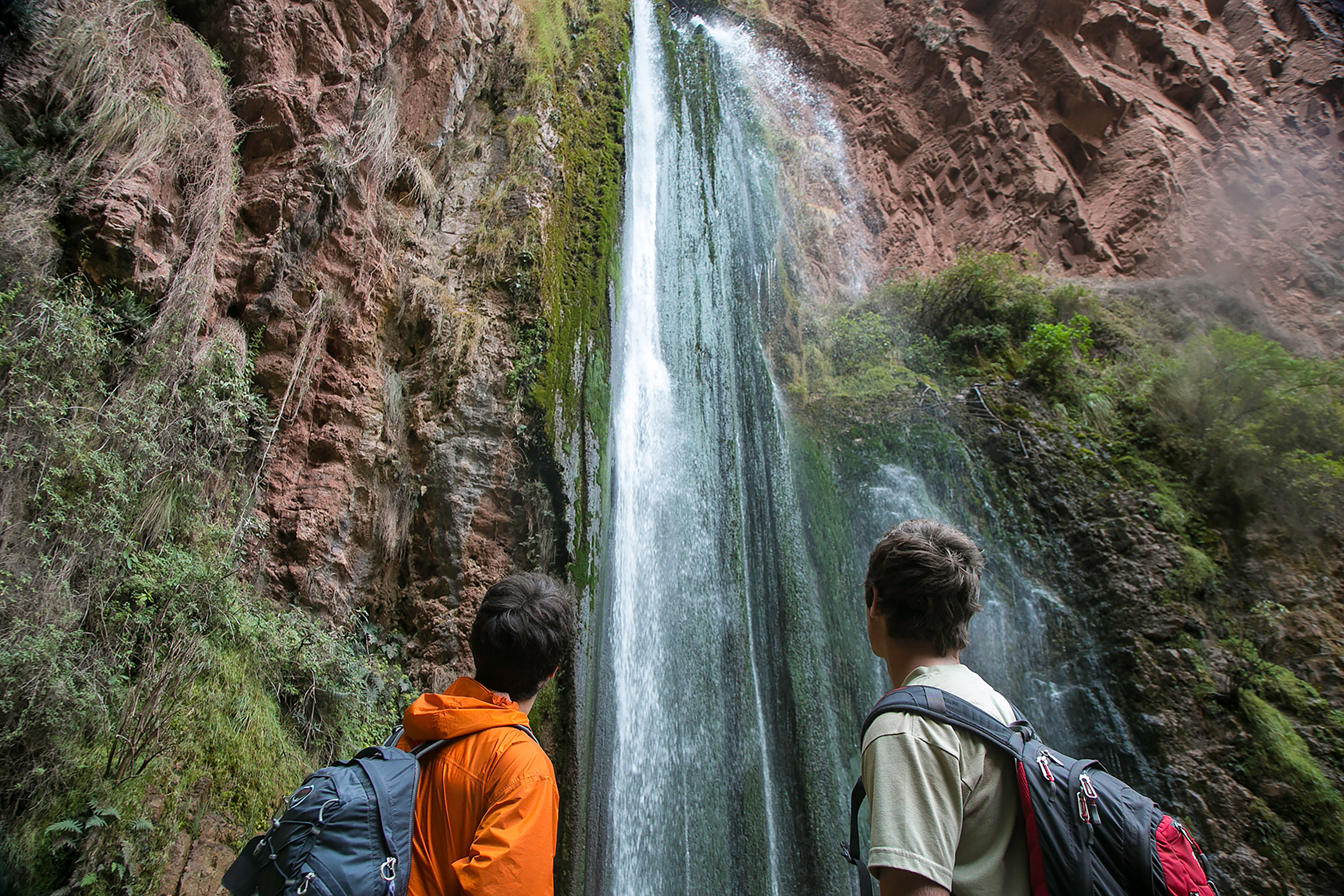 Student travelers with backpacks gaze up at a tall waterfall cascading down a rocky cliff
