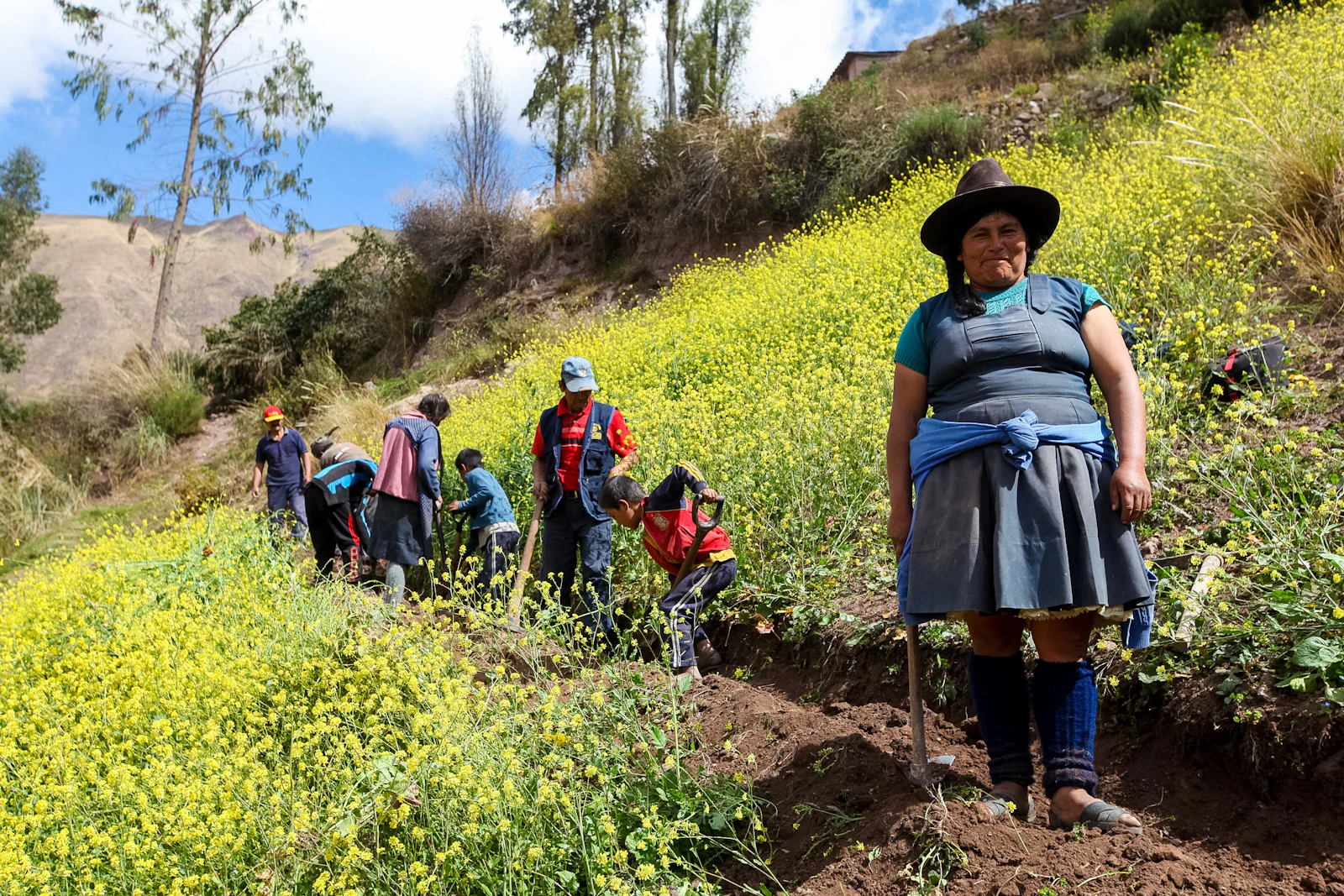 A local woman in a wide-brimmed hat stands on a flowering hillside as teen travelers dig a trench below her in Peru