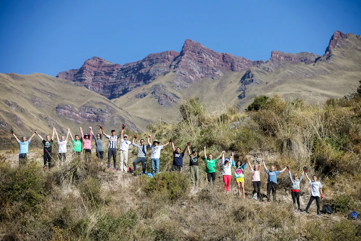 A large group of teen travelers raise their arms in a line on a scrubby hillside beneath jagged Andean peaks in Peru