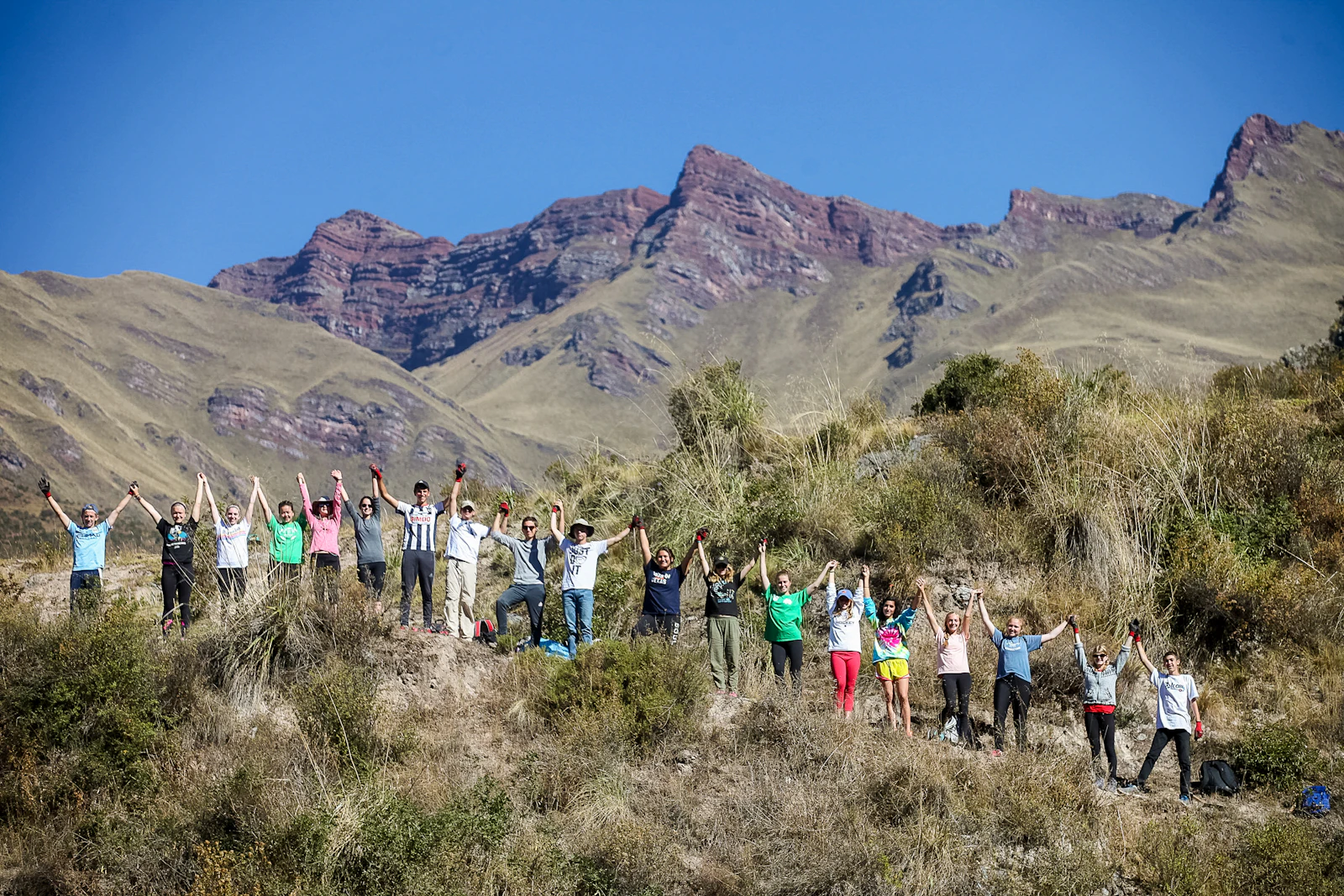A large group of teen travelers raise their arms in a line on a scrubby hillside beneath jagged Andean peaks in Peru