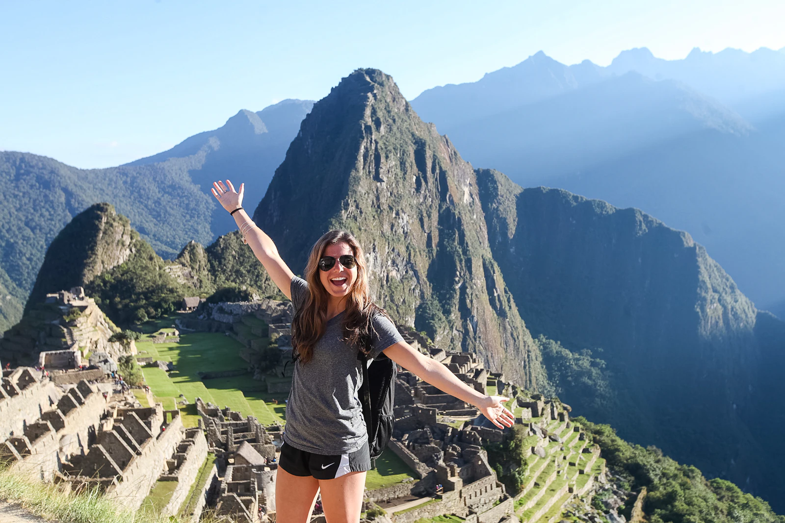 A teen traveler in sunglasses throws her arms wide above the ruins of Machu Picchu with Huayna Picchu behind