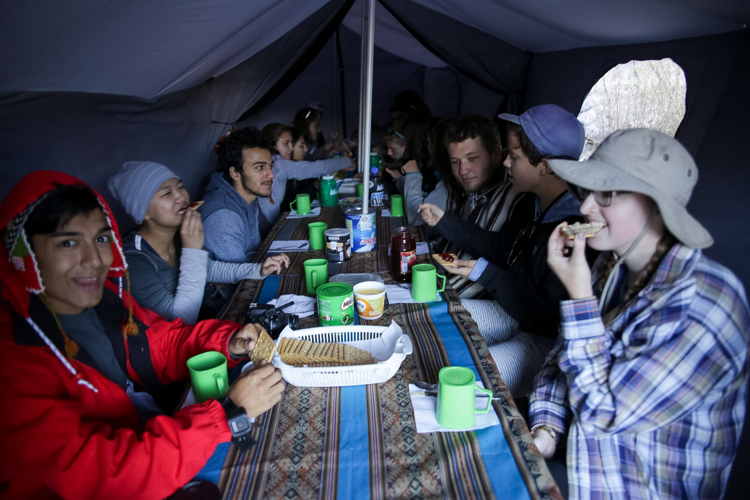 Students share a meal at a long table inside a dining tent, mugs and bread laid out