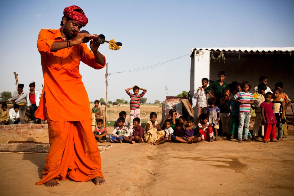 A performer in an orange turban and robe plays a flute for a crowd of onlookers