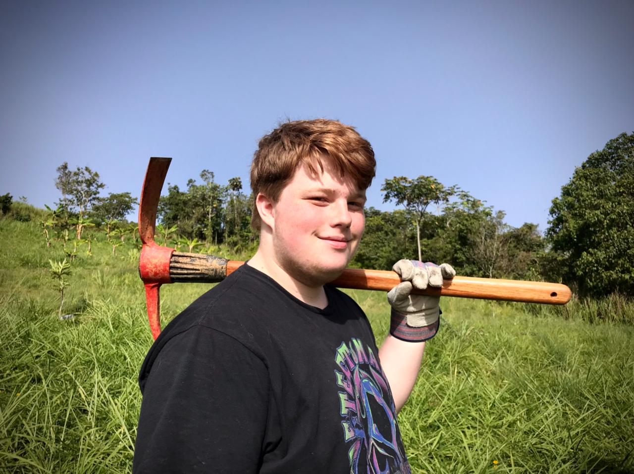 A student carries a farming tool over his shoulder in a grassy field