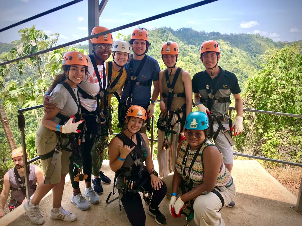 Teens in helmets and zipline harnesses pose on a platform above a forested valley