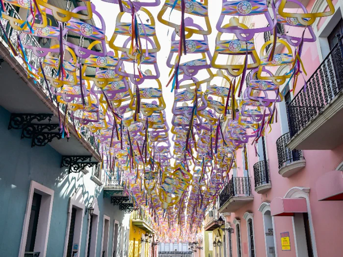 A cobbled Old San Juan lane under a canopy of hanging paper decorations, lined with pink and blue balconied buildings.