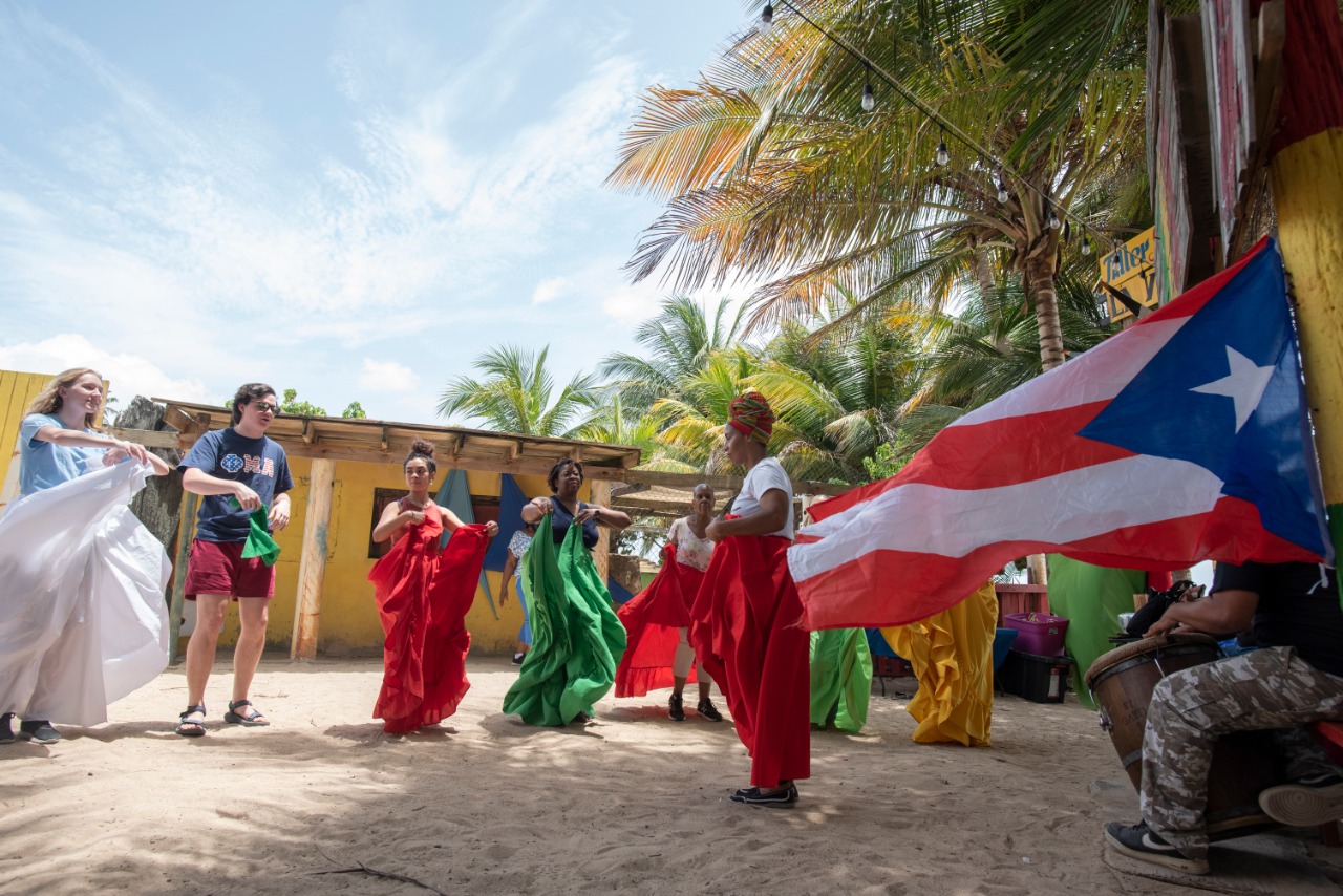 Students in traditional dress dance alongside a large Puerto Rican flag during an outdoor cultural performance