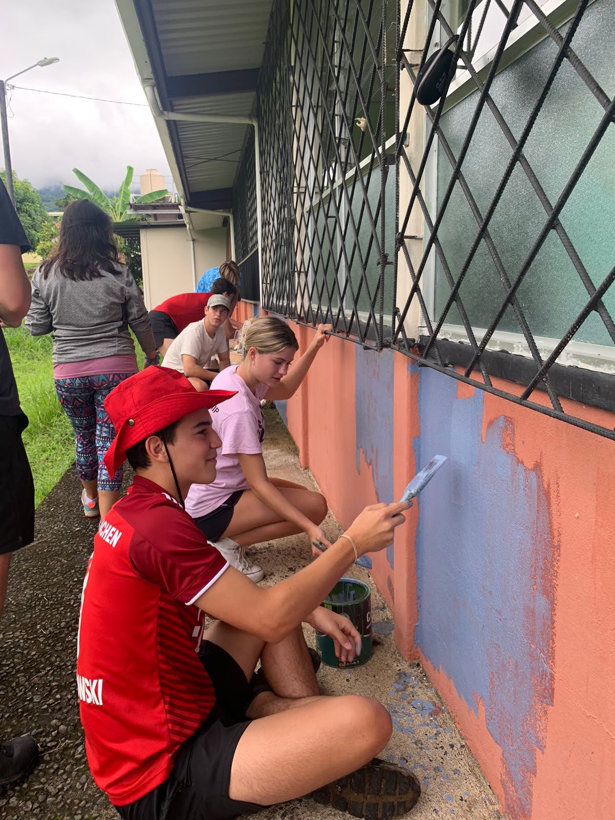 PVS School service. Photo of students during service painting an orange house in a more blue/gray color. Sitting down and painting with the student in front wearing a read outfit with a read, matching hat.
