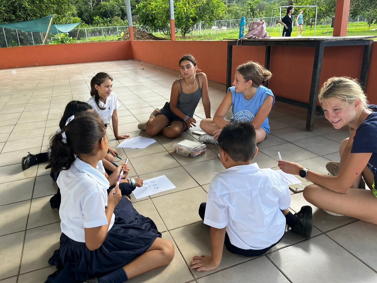 Students during PVS School service activity sitting down outside on the school. They sit in a circle with their uniforms, with our Rustic Pathways students. They have papers in front of them on the ground and seem to be discussing or learning something together. 7 people all together.