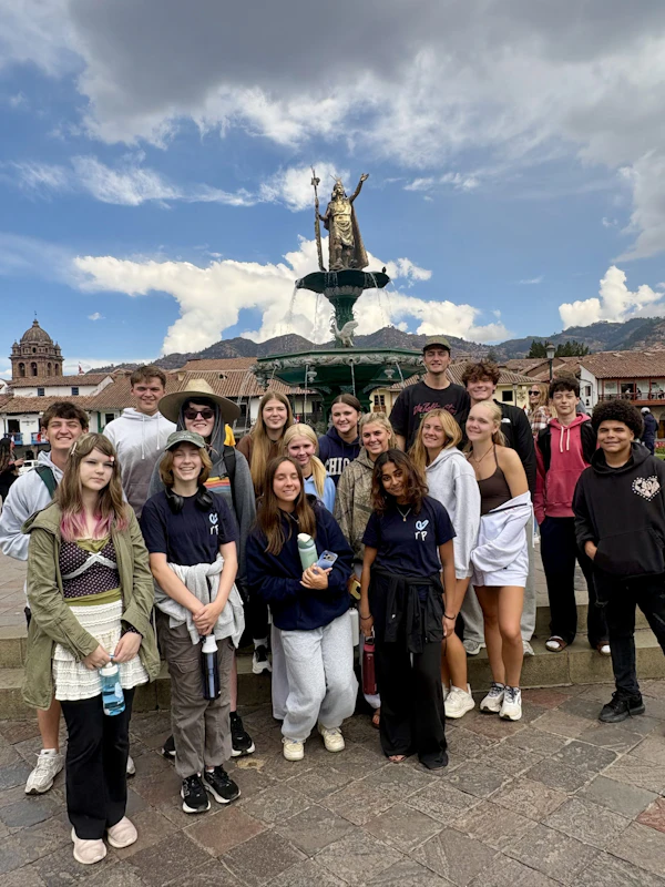 Teen travelers gather around the Pachacutec fountain in the Plaza de Armas in Cusco, with colonial rooftops and the Andes behind