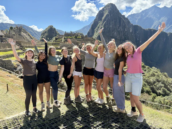 Eleven teen travelers stand in a line with their arms raised on a terrace at Machu Picchu, with Huayna Picchu rising behind them