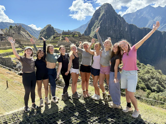 Eleven teen travelers stand in a line with their arms raised on a terrace at Machu Picchu, with Huayna Picchu rising behind them.