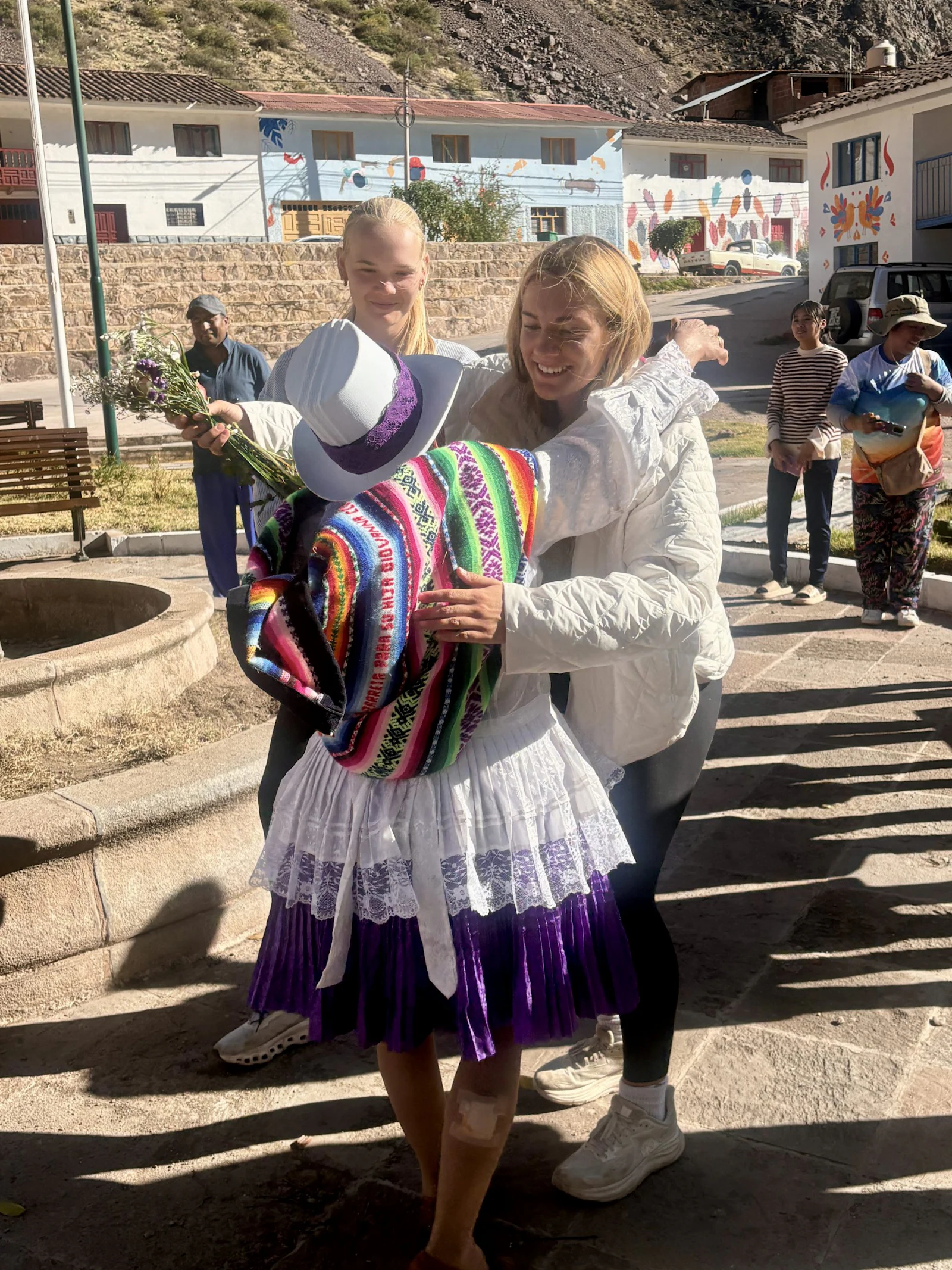 Two teen travelers hug a host in traditional Andean dress, with a layered skirt and white hat, in a village square in Peru