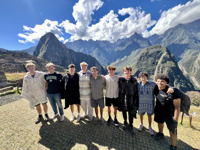 Nine teen travelers in woven ponchos and jackets stand together on a terrace at Machu Picchu with the ridgeline of the Andes behind.