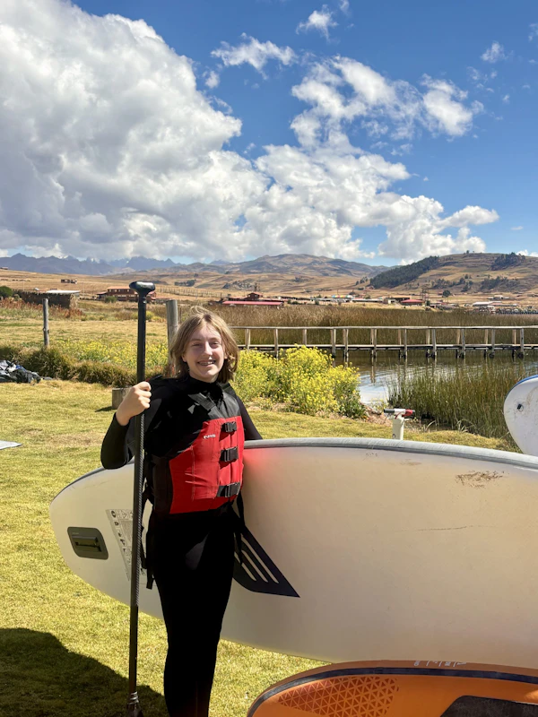 A teen traveler in a wetsuit and red buoyancy aid holds a paddleboard and paddle on the grass beside a highland lagoon in Peru