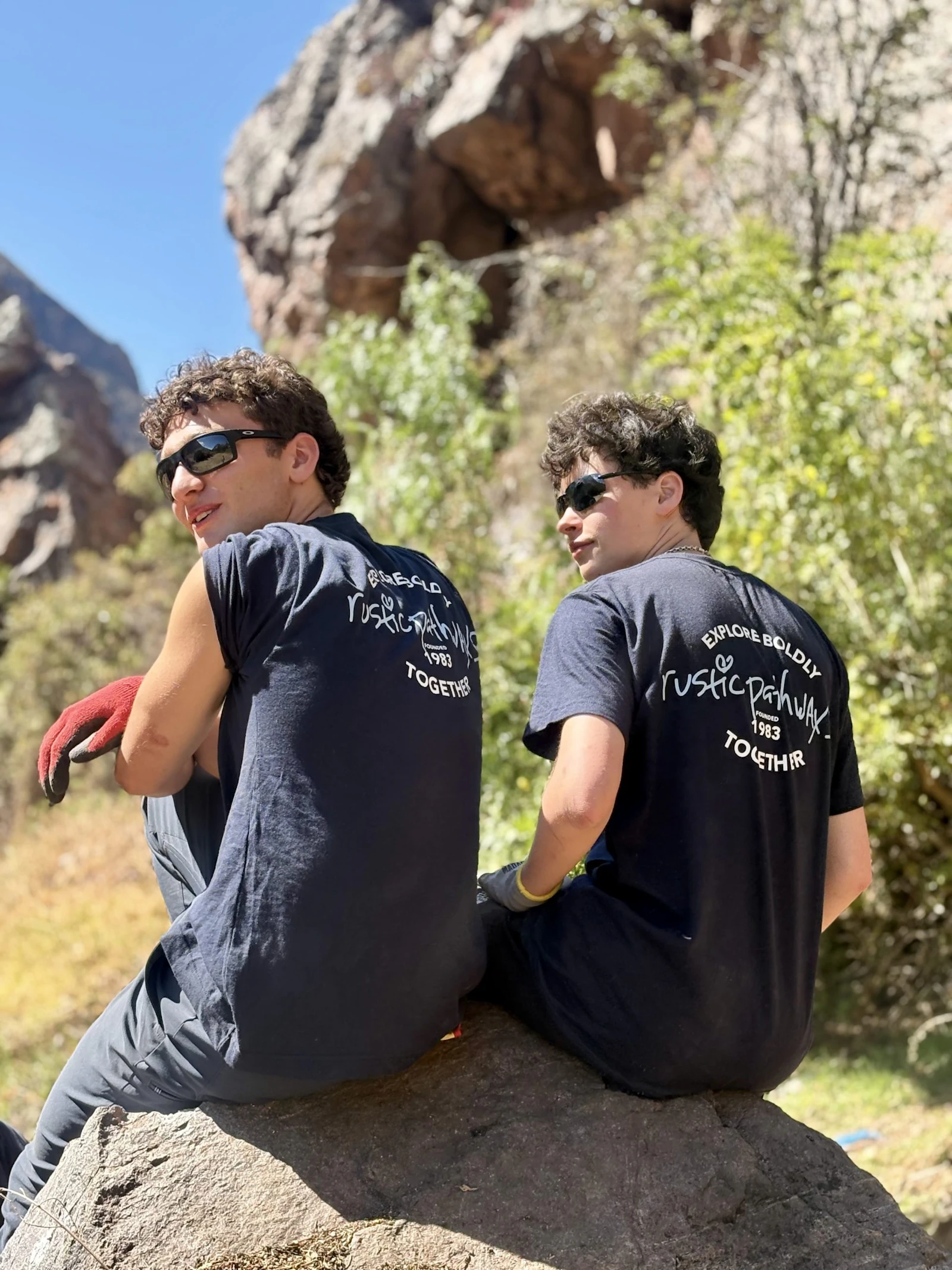 Two teen travelers in Rustic Pathways shirts sit on a large boulder below a rocky, wooded slope