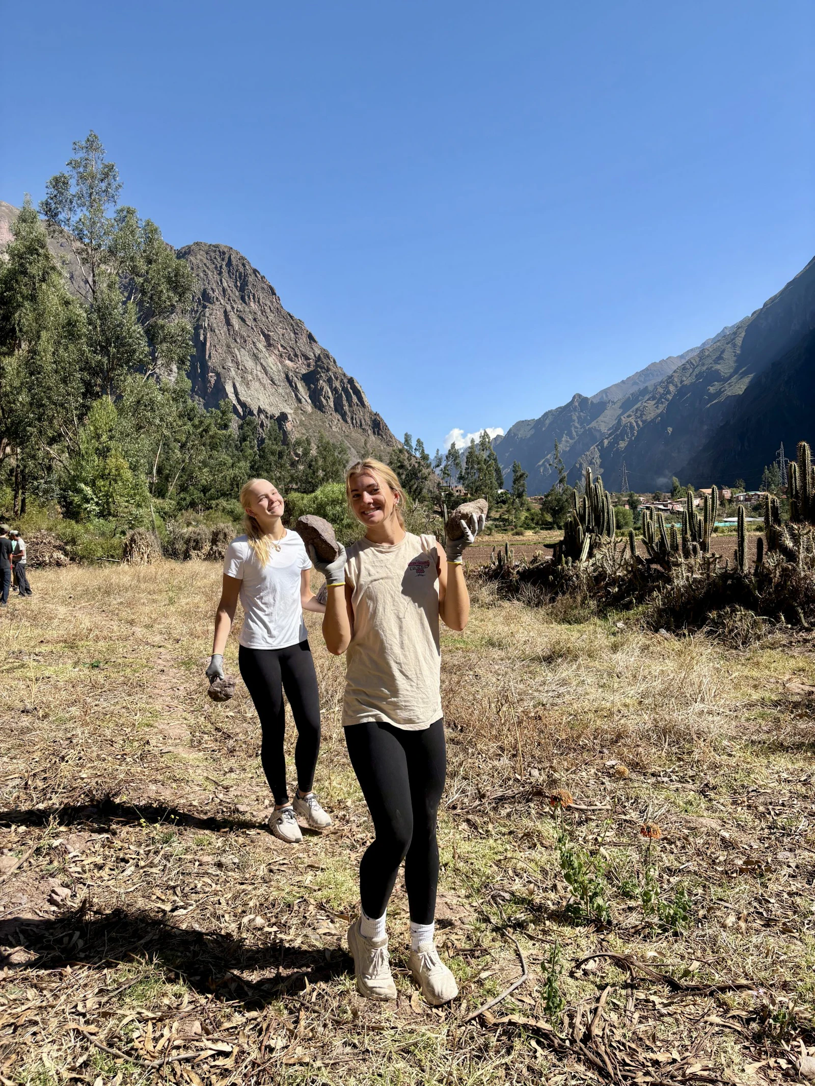 Two teen travelers in work gloves carry rocks across a cleared valley floor in Peru, with cactus, eucalyptus and mountain peaks behind