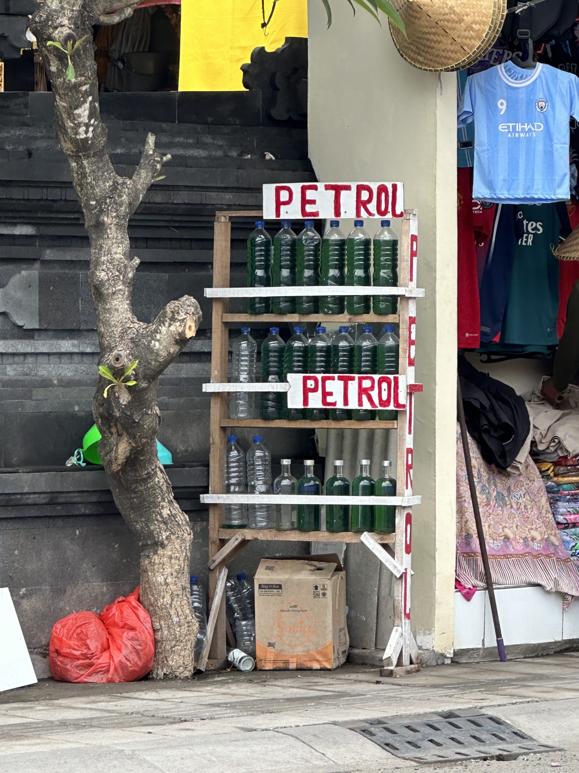 A roadside stall selling bottled petrol in Bali, displayed in green bottles on wooden shelves.