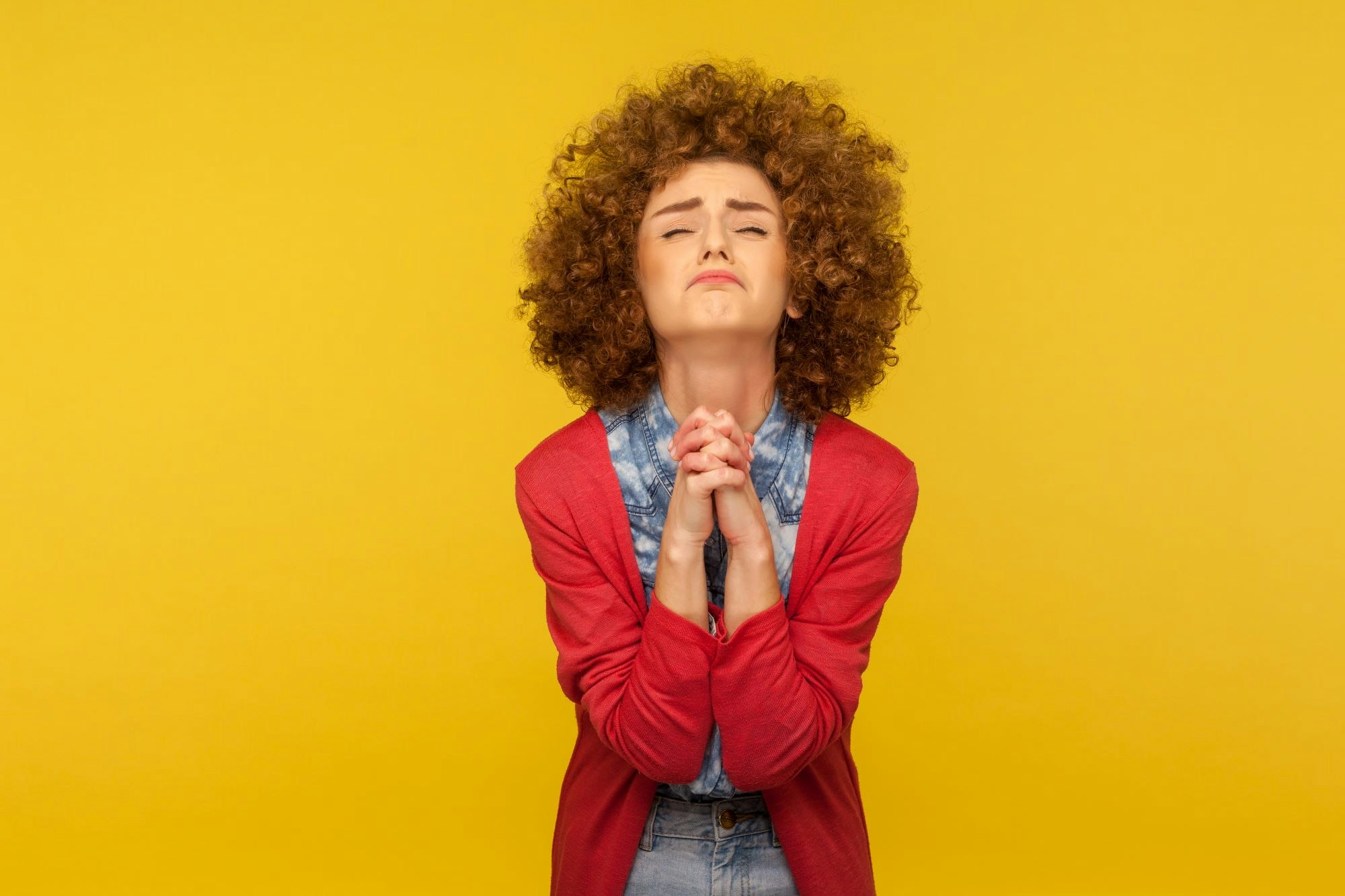A curly-haired woman in a red cardigan clasps her hands together in a pleading gesture against a bright yellow background.