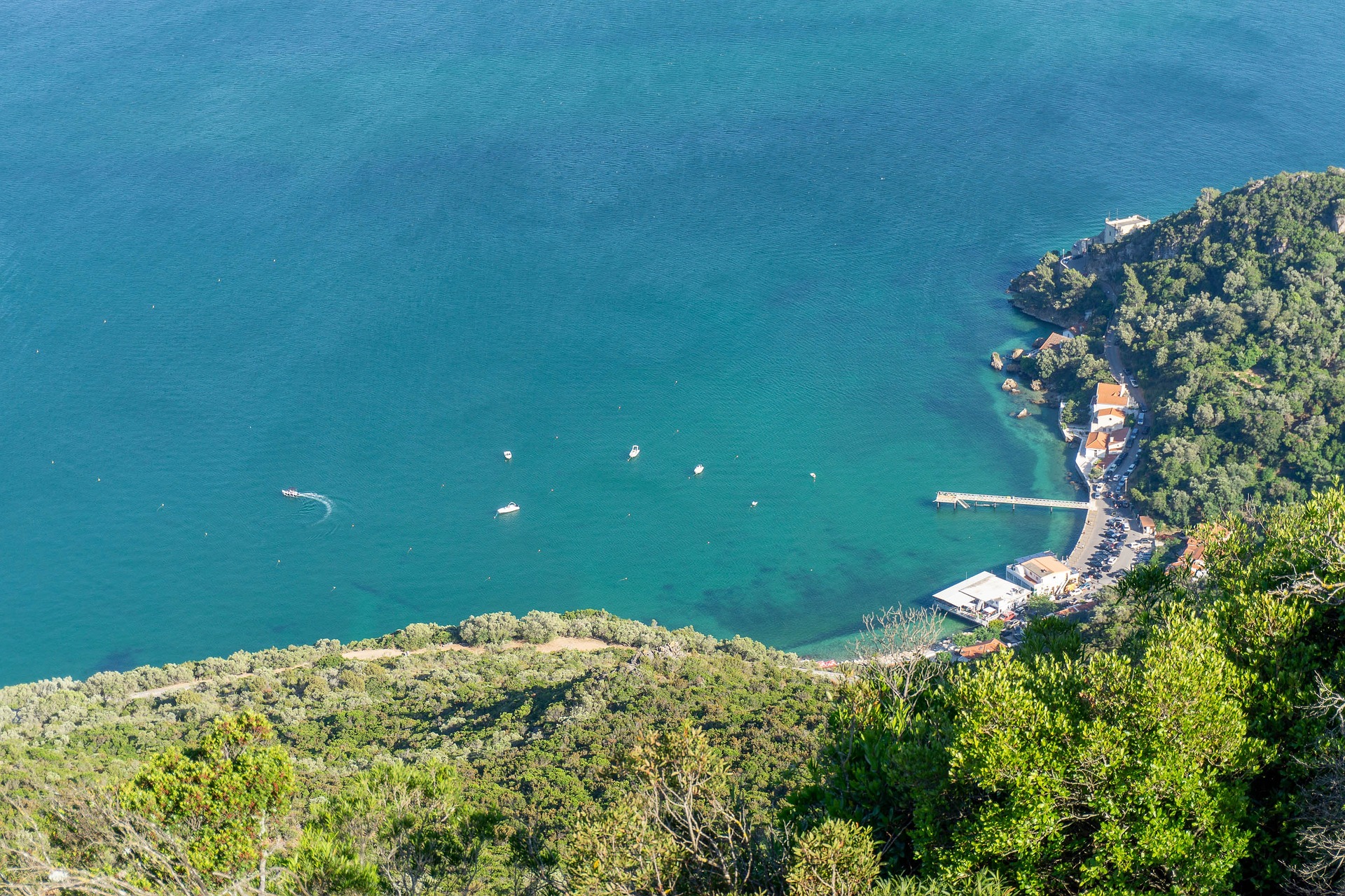 A sheltered turquoise bay seen from a wooded ridge, with small boats moored off a short jetty and a strip of low buildings at the water's edge.