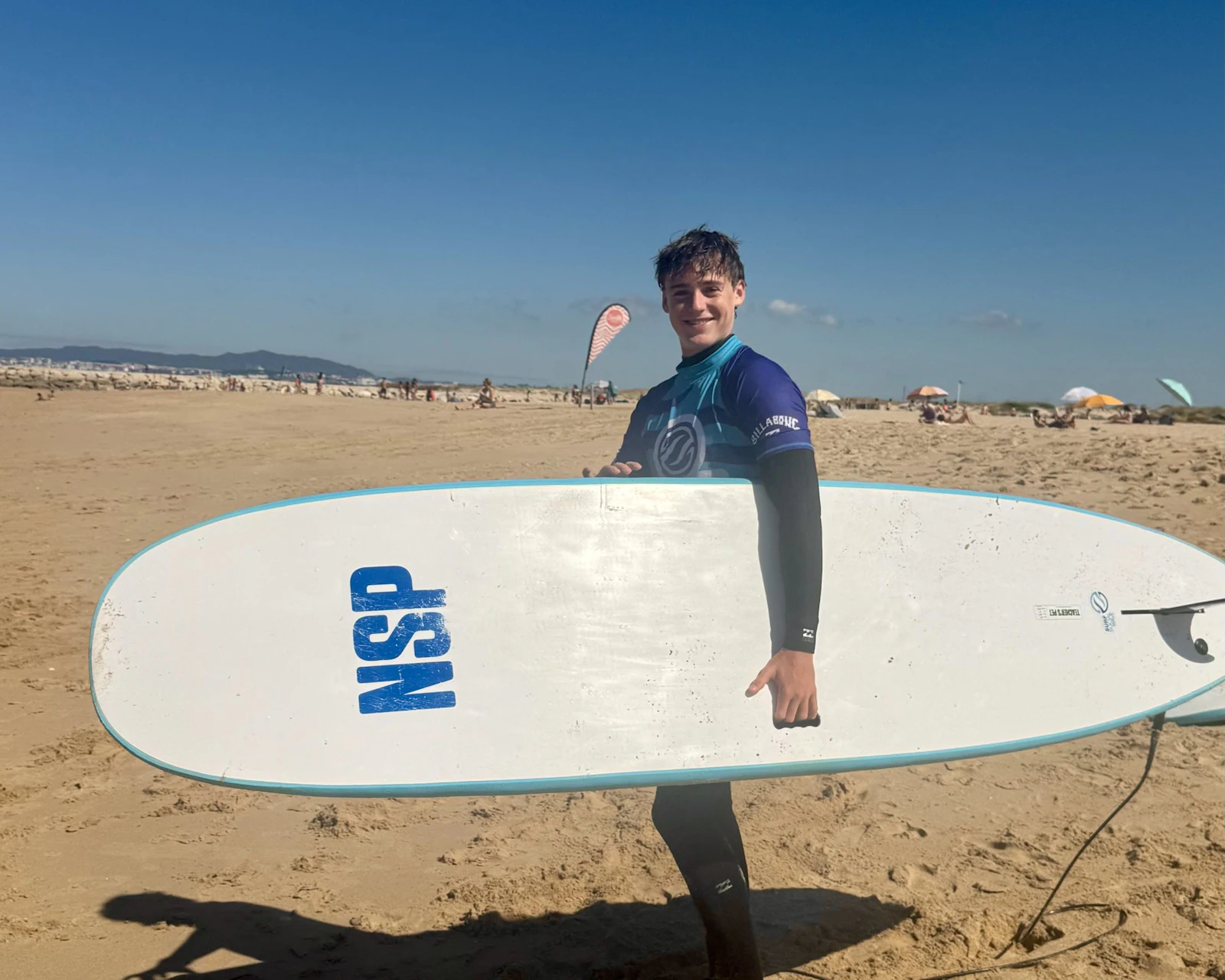 A teen traveler in a wetsuit carries a white surfboard under one arm on a wide sandy beach in Portugal