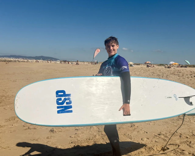 A teen traveler in a wetsuit carries a white surfboard across a wide sandy beach in Portugal