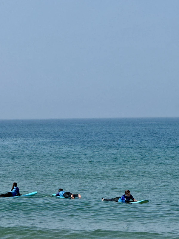 Three teen travelers in wetsuits paddle soft-top surfboards in calm water off a beach in Portugal