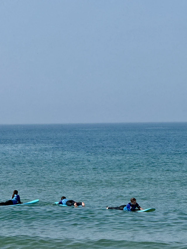 Three teen travelers in wetsuits paddle soft-top surfboards in calm water off a beach in Portugal