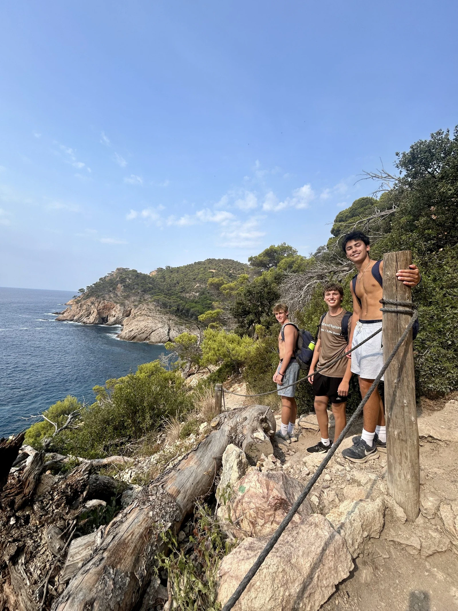 Three teen travelers stand on a rocky coastal path in Spain above a blue cove, with pines growing from the headland behind