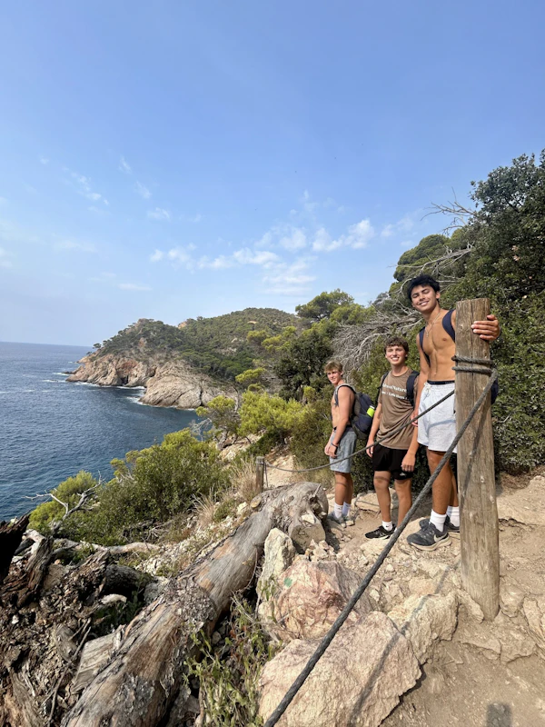 Three teen travelers stand on a rocky coastal path above a blue cove, with pines growing from the headland behind them