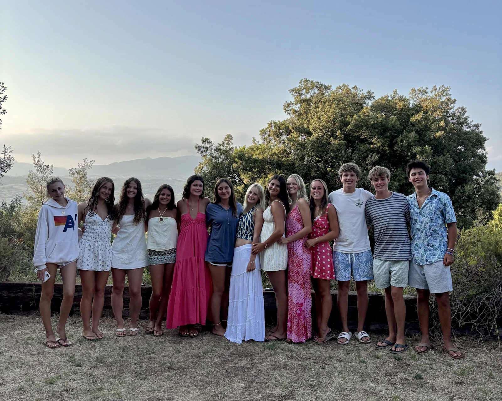 Thirteen teen travelers stand in a line on a grassy hilltop at dusk with a wide valley and a hazy ridgeline behind them