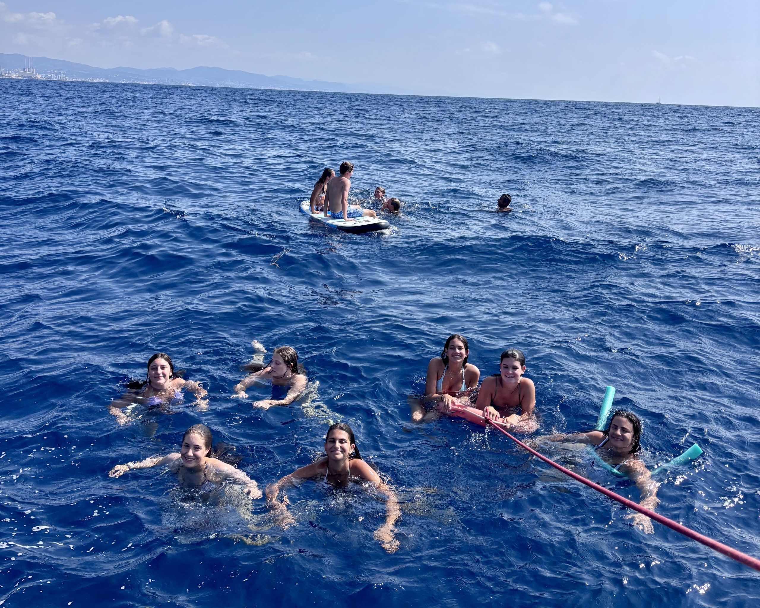 Eight teen travelers swim in deep blue Mediterranean water off Spain beside a paddleboard and a floating line, with the coastline in the distance