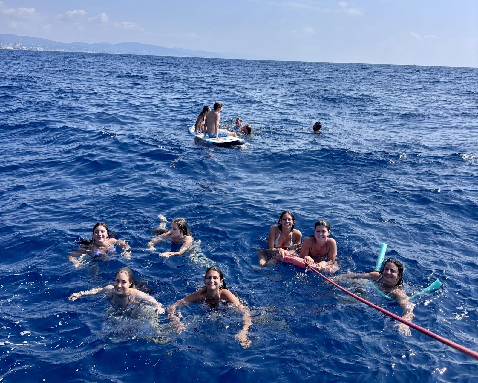 Eight teen travelers swim in deep blue sea water beside a paddleboard and a floating line, the coast far behind them
