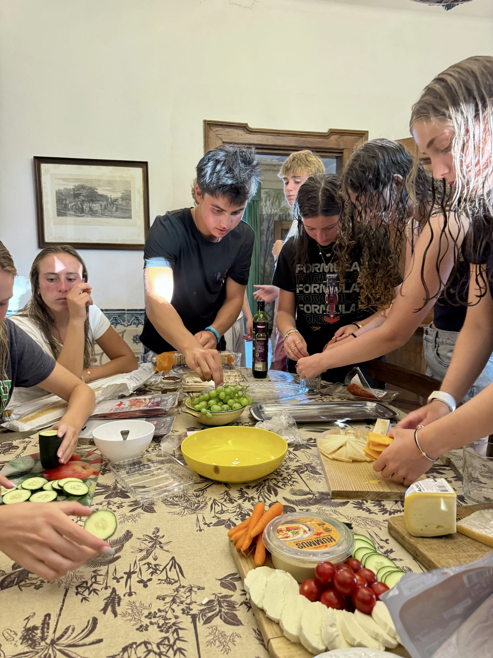 Teen travelers crowd around a long table slicing bread and cheese and preparing vegetables together