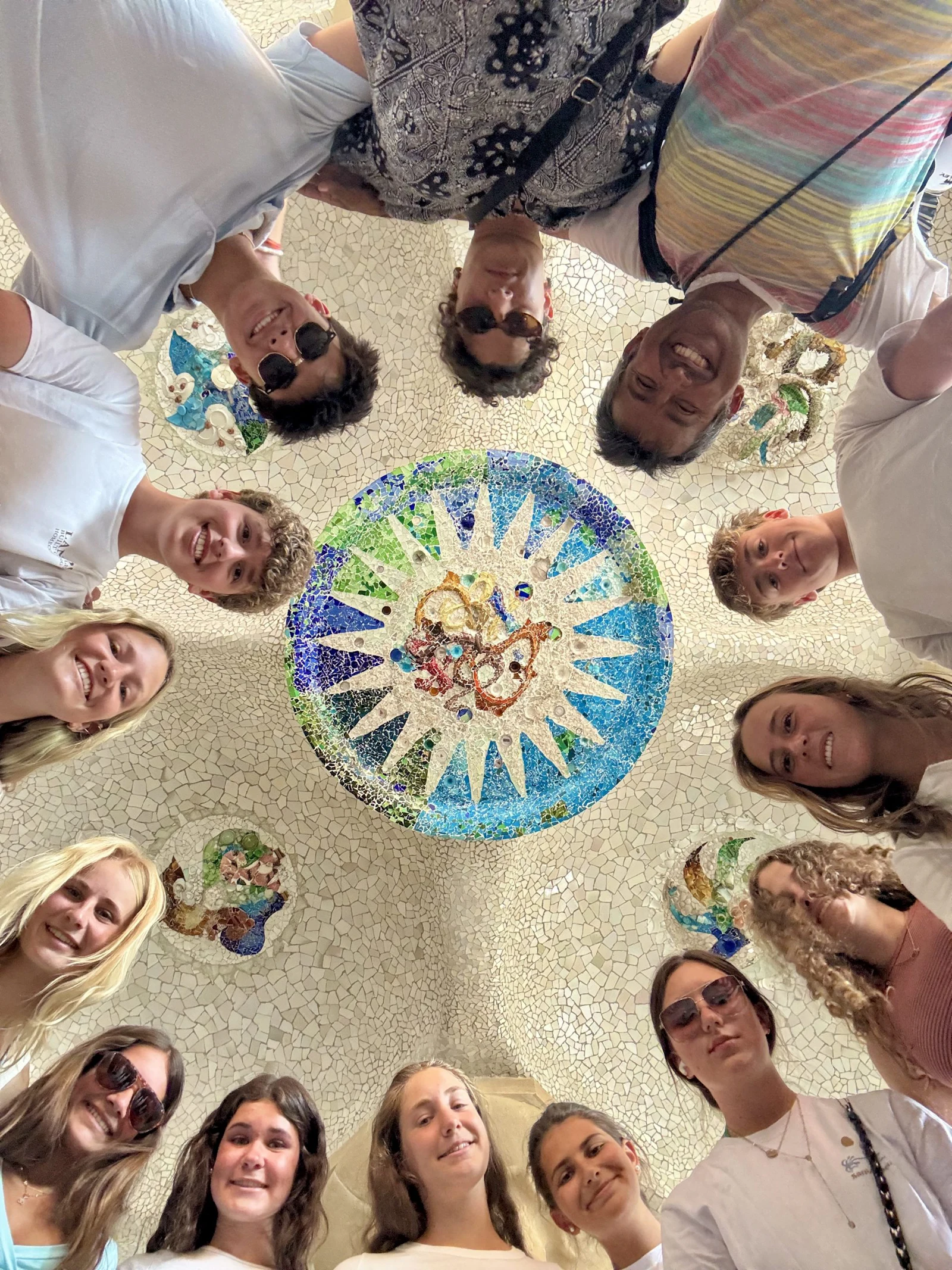 Teen travelers stand in a ring under a round mosaic medallion in the ceiling of Park Güell's Hypostyle Hall in Barcelona