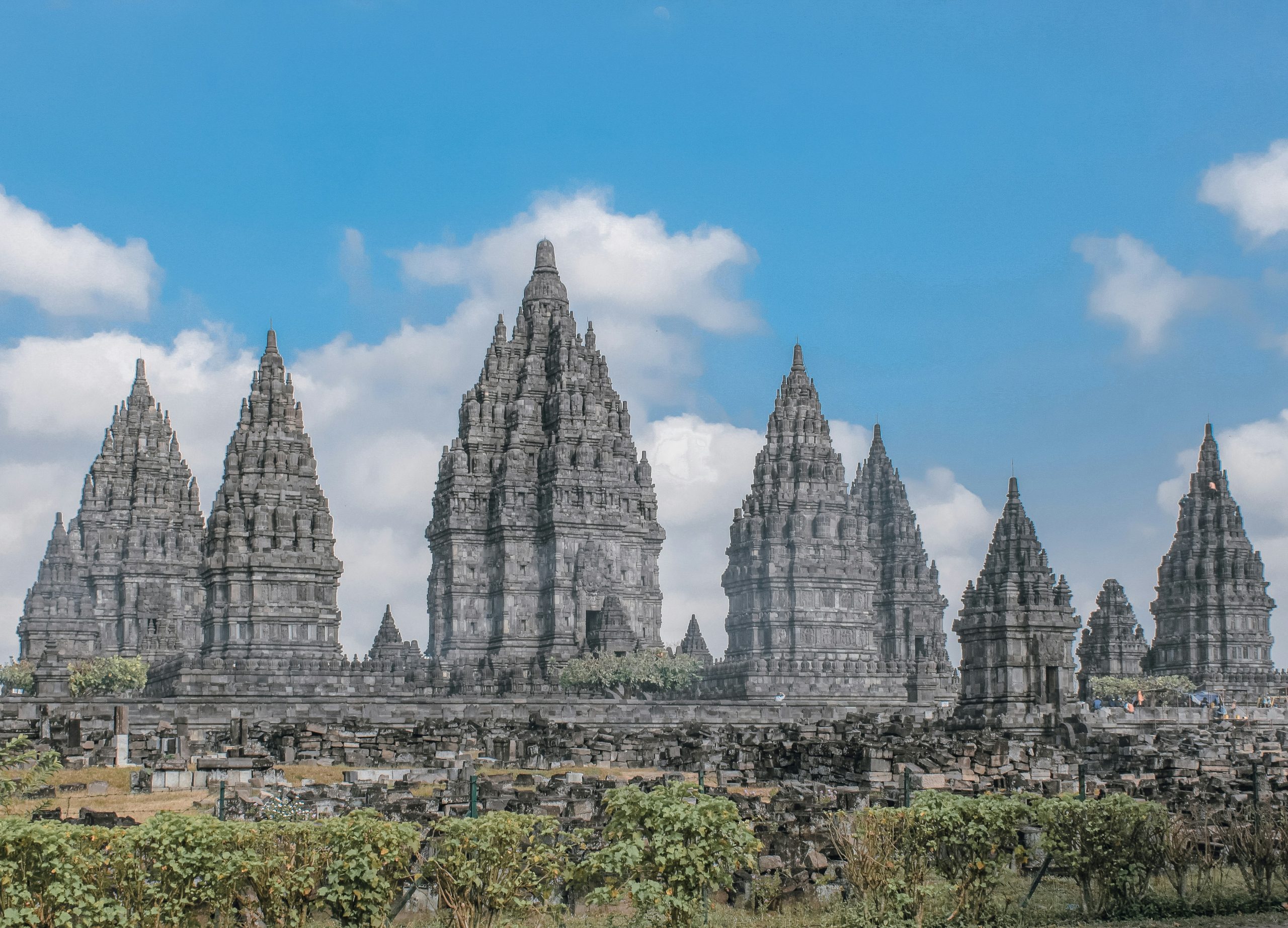 Rows of tall, pointed stone temple towers rise against a partly cloudy blue sky at Prambanan.
