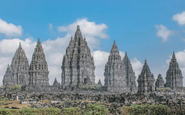 Rows of tall, pointed stone temple towers rise against a partly cloudy blue sky at Prambanan.