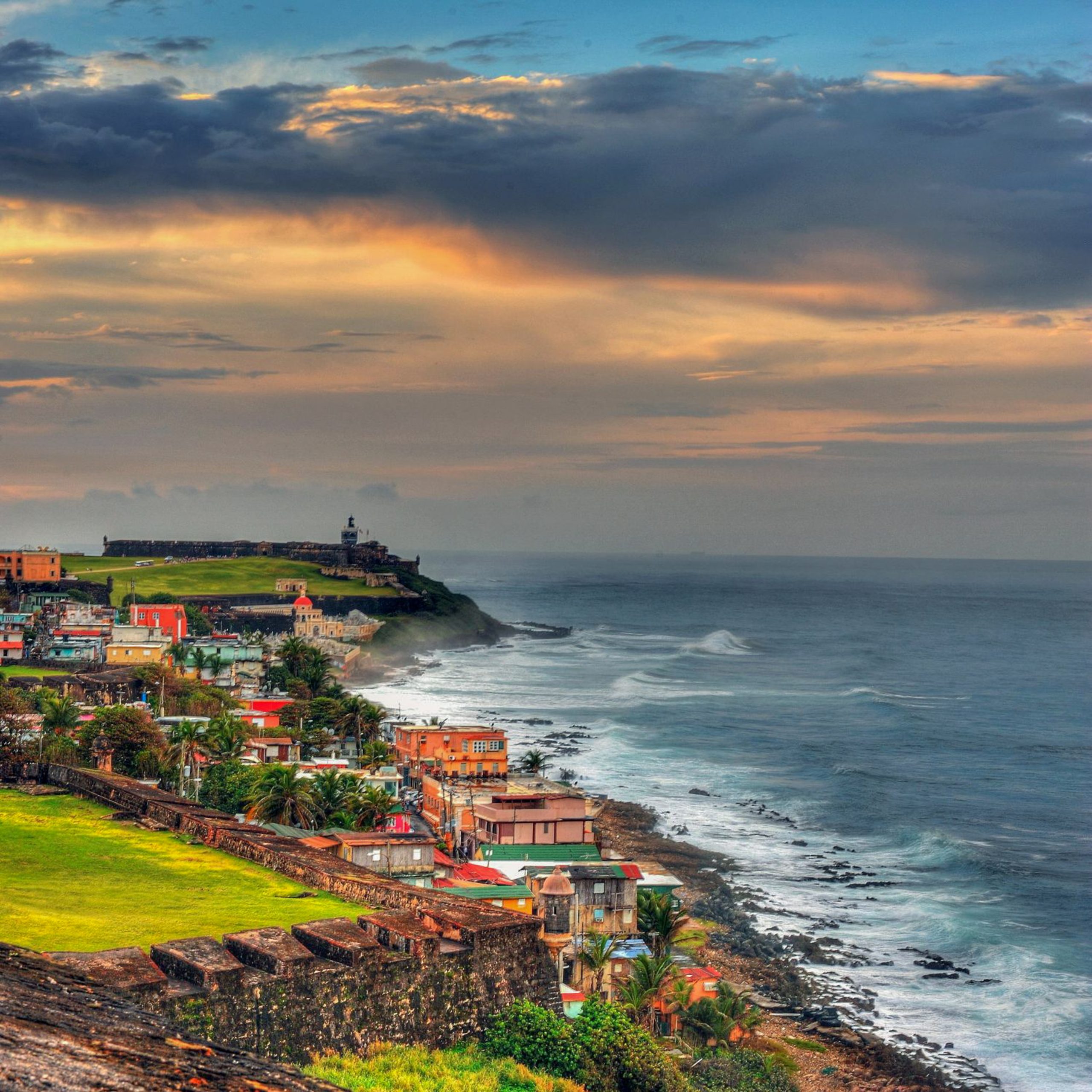 A coastal city in Puerto Rico hugs the cliffside above crashing waves under a sunset sky