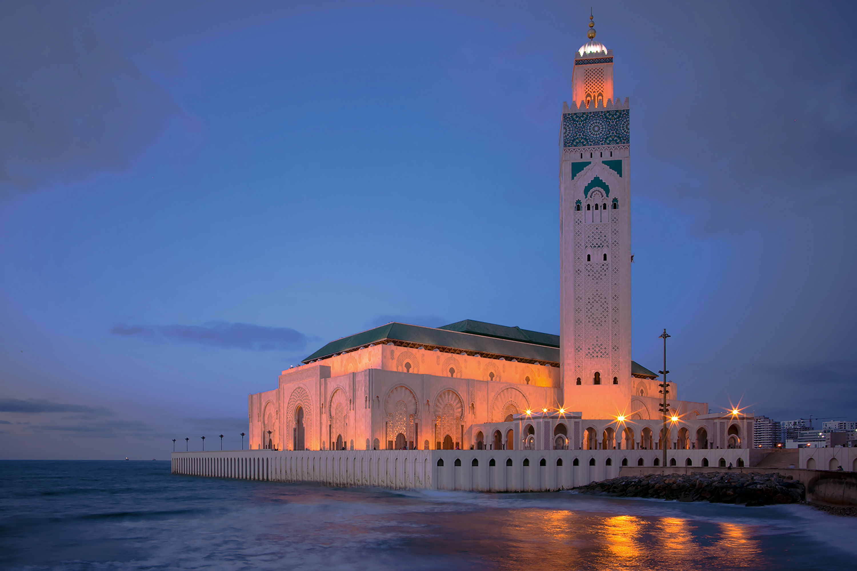 A grand mosque with a tall minaret illuminated at dusk beside the ocean