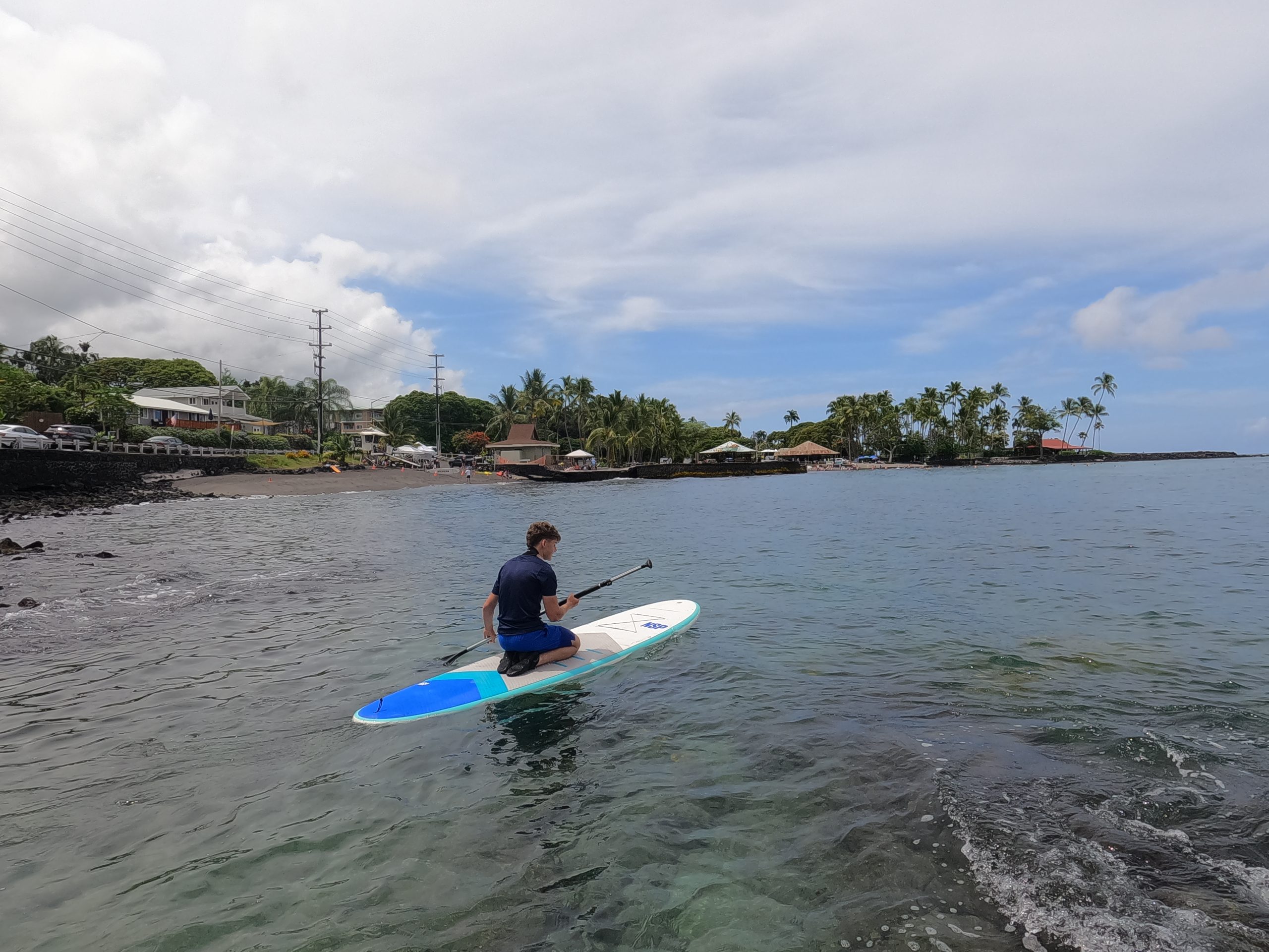 A student kneels on a paddleboard near a palm-lined shoreline