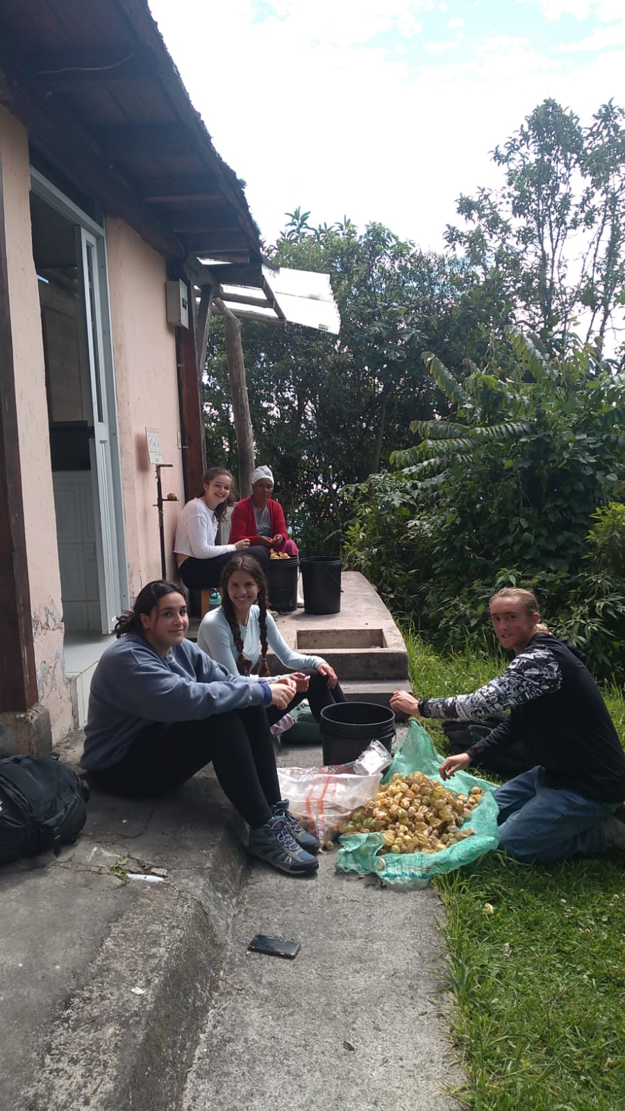 Students sit on the front steps of a house sorting produce into a bucket alongside a local host.