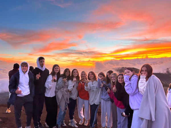 A group of teen travelers in hoodies and blankets stands above a sea of clouds under an orange sunset sky