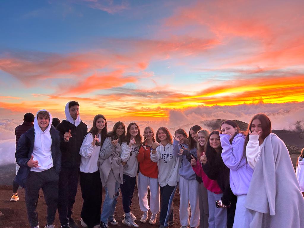 Students give a thumbs-up on a mountainside as clouds glow under a colorful sunset sky