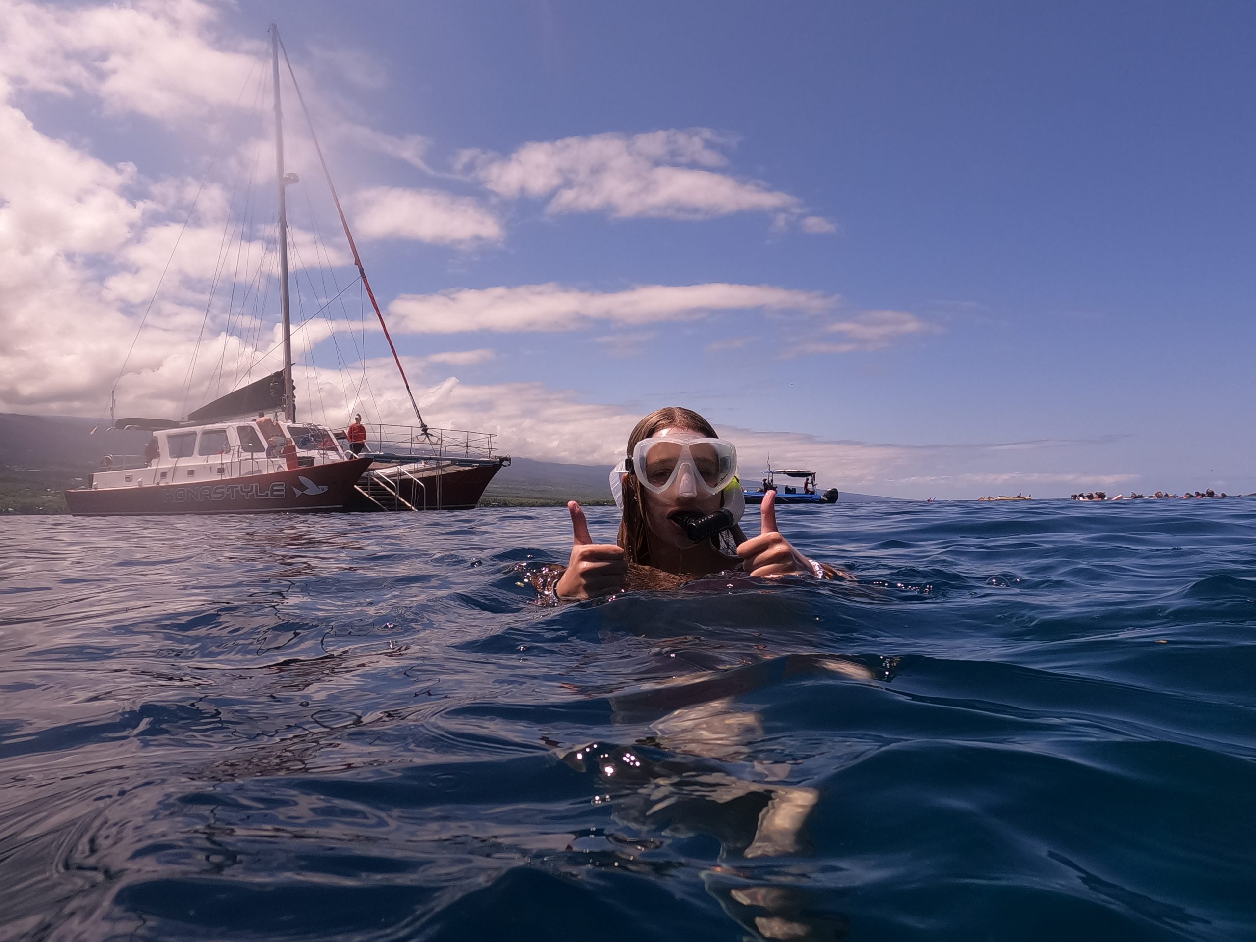 On a school trip, a student gives thumbs up while snorkeling near a sailboat off the Kona coast