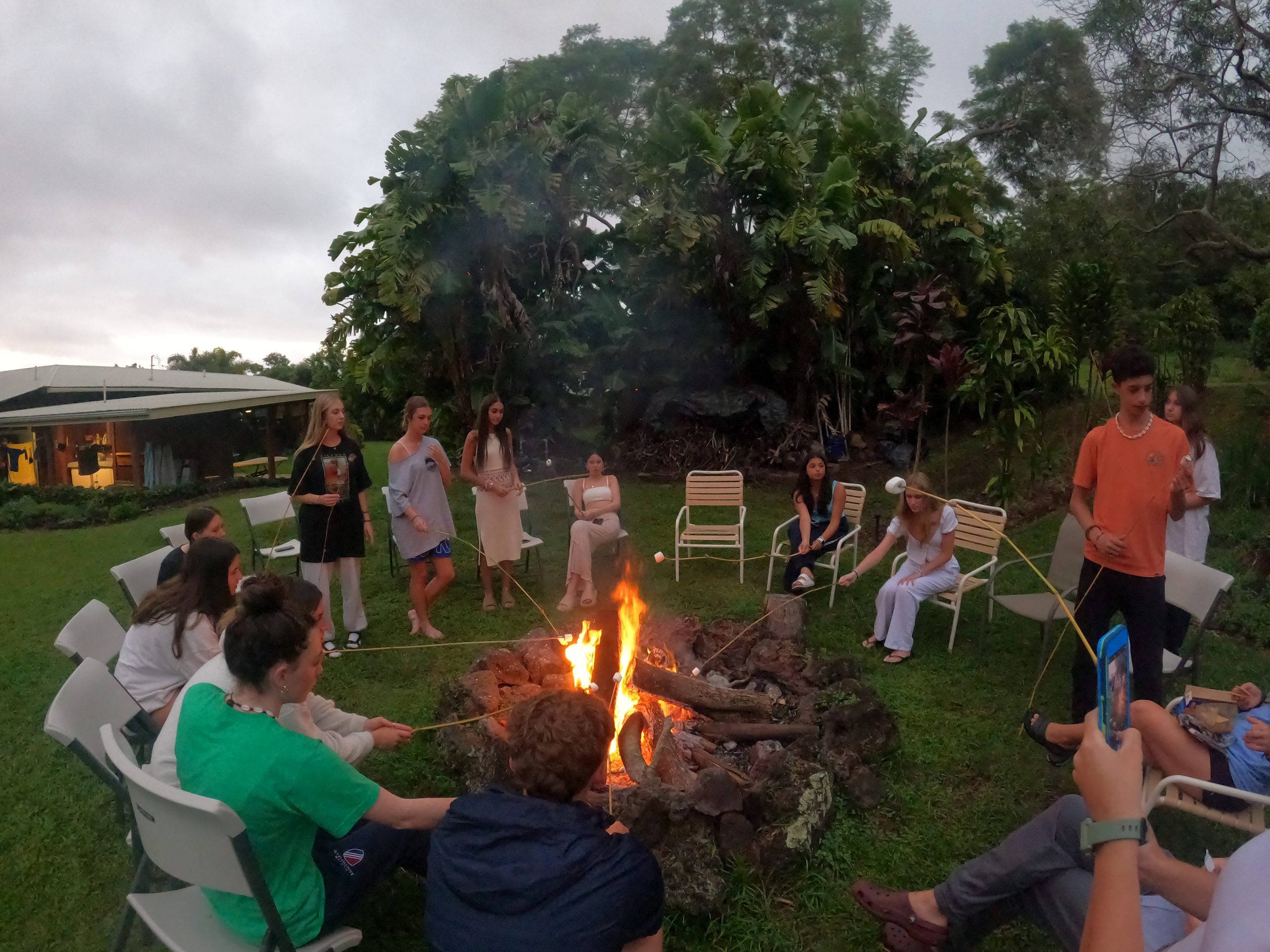 Students gather around a bonfire at dusk, roasting marshmallows on sticks